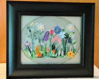 May include: A framed paper quilling art piece featuring a colourful garden scene with various flowers and green foliage. The artwork is enclosed in a black rectangular frame, showcasing intricate coiled paper strips in shades of blue, pink, orange, purple, and white against a light grey oval background.