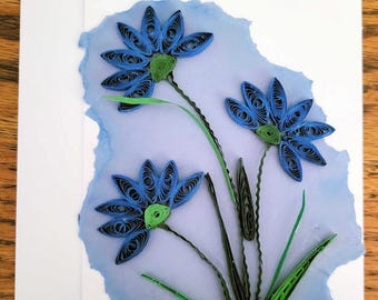 May include: A white greeting card featuring a paper quilling design of three blue flowers with green centres and stems. The flowers are arranged on a textured, light blue painted background with torn edges, creating a layered, dimensional effect.