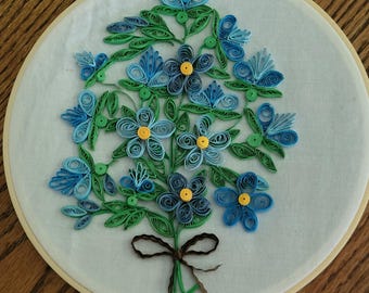 May include: A paper quilling art piece inside a circular wooden embroidery hoop. The design features a bouquet of blue flowers with yellow centres and green leaves made from coiled paper strips, tied together with a brown paper ribbon.