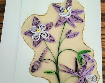 May include: A greeting card featuring a paper quilling design of three purple and white flowers with yellow centres. The floral arrangement includes green stems and leaves, all mounted on a light beige, irregularly shaped paper cutout.