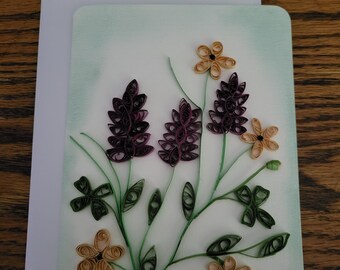 May include: A rectangular greeting card featuring a paper quilling design of purple lupine-like flowers, light tan five-petaled blossoms, and green stems with leaves. The artwork is set against a white background with a soft, light green airbrushed border.