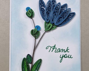 May include: A greeting card featuring a paper quilling design of a blue flower with green leaves and stems on a light blue background. The text "Thank you" is written in green cursive script on the lower right side of the card surface.