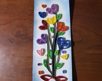 May include: A rectangular paper bookmark with a light blue watercolour-style background and a blue ribbon at the top. The design features a quilled paper bouquet with colourful hearts in purple, yellow, orange, red, pink, and blue, along with green leaves and a red quilled flower at the base.