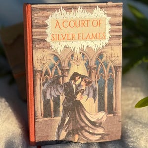 Puede incluir: Un libro de tapa dura titulado A Court of Silver Flames con una cubierta de tela texturizada. La portada presenta una ilustración de dos figuras bailando frente a ventanas arqueadas, coronada con un gráfico de llamas. El lomo es naranja con un marcapáginas rojo.