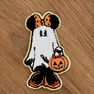 May include: An embroidered patch featuring a character dressed as a ghost, wearing black ears with an orange polka-dot bow, a white sheet, and black shoes with orange accents. The character holds an orange jack-o'-lantern bucket with a gold border.