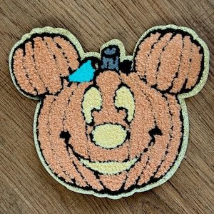May include: A textured, punch needle patch shaped like a pumpkin with mouse-like ears. The pumpkin is orange with a light yellow face, a small blue bow, and a dark stem. The entire patch is outlined with a shimmering gold glitter border.
