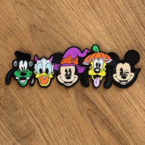 Op de afbeelding: Een set van vijf geborduurde patches met Disney-figuren in Halloween-kostuums. De ontwerpen bevatten Goofy als Frankenstein, Donald Duck met paarse hoorns, Minnie Mouse met een paarse heksenhoed, Pluto met een pompoenhoed en Mickey Mouse.