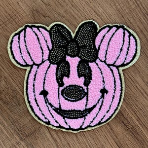 Op de afbeelding: Een patch in de vorm van een pompoen met muizenoortjes, voorzien van een roze textuur, een zwarte glitterstrik, zwarte glitterogen, een zwarte glitterneus en een zwarte glittermond, omlijnd met een glinsterende gouden glitterrand.