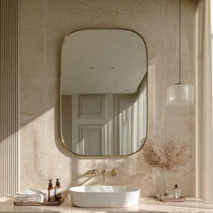 May include: A rectangular wall mirror with rounded corners and a thin gold frame mounted above a white oval vessel sink. The sink features a gold faucet and is set on a beige marble countertop with a stack of white towels and a glass vase.