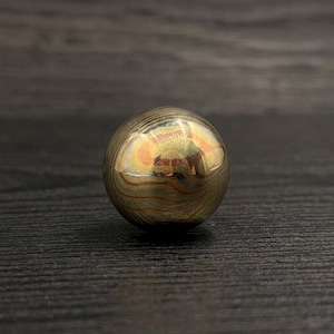 May include: A polished spherical object featuring a layered, swirling pattern of metallic gold, bronze, and dark brown tones. The surface is highly reflective, showing distorted light reflections across its smooth, rounded, and continuous exterior.