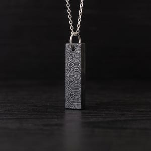 May include: A rectangular pendant necklace featuring a dark gray metal bar with a complex, swirling Damascus steel pattern. The pendant hangs from a thin, silver-colored metal chain that attaches through a circular jump ring at the top center.