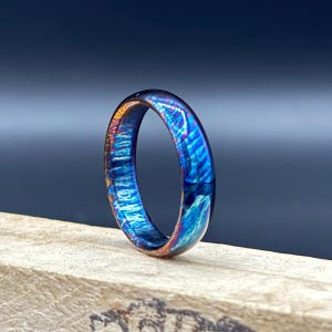 May include: A ring featuring a vibrant, iridescent finish with swirling patterns of deep blue, purple, and copper tones. The band has a smooth, polished surface that reflects light, creating a dynamic color shift across its circular structure.