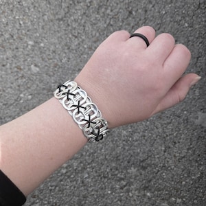 May include: A bracelet constructed from silver-colored metal soda can pull tabs linked together with black cord in a crisscross pattern, accompanied by a simple black ring worn on the finger. The design features a repeating circular tab structure.