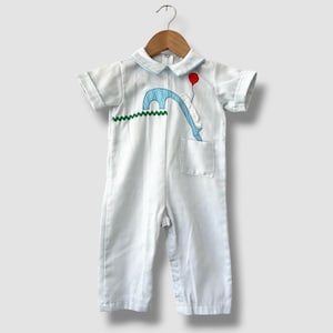 May include: A white and light blue pinstriped short-sleeve romper featuring a Peter Pan collar, a green zigzag trim, and an appliqué of a blue polka-dot elephant holding a red balloon. The garment includes a small chest pocket and button closures.