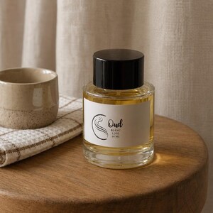 May include: A clear glass perfume bottle with a black cylindrical cap and a white label. The label features a black swirl logo and the text Oud BLANC 1.7OZ 50 ML. The bottle contains a golden liquid and sits on a wooden surface next to a small cup.