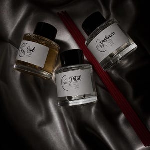 May include: Three clear glass diffuser bottles with black caps and white labels. The labels read Oud Blanc, Petal, and Cashmere Cocoa. Each bottle contains 1.7 oz and 50 ml of liquid. A set of long, thin, dark red reed sticks lies beside the bottles.