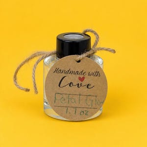 May include: A clear glass bottle with a black cap, containing a transparent liquid. A round brown paper tag is tied to the neck with twine, featuring the printed text "Handmade with Love" and handwritten text "Petal Glow" and "1.7 oz".