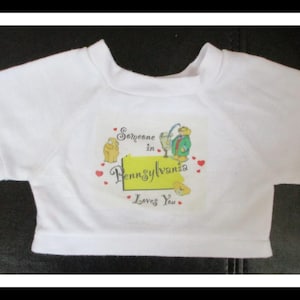 Könnte beinhalten: Ein weißes kurzärmeliges T-Shirt mit einem Grafikdruck, der eine gelbe Karte von Pennsylvania, kleine Teddybären, rote Herzen und den Text Someone in Pennsylvania Loves You in schwarzer und blauer Schrift auf der Brust zeigt.