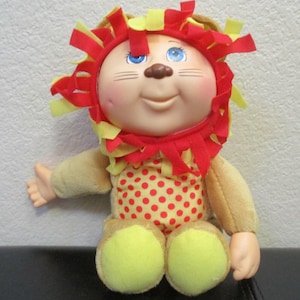 May include: A plush doll with a vinyl face, blue eyes, and a brown nose. The doll wears a lion-themed hood featuring red and yellow fabric strips as a mane. The body is tan with a red and yellow polka-dot pattern on the chest and yellow feet.