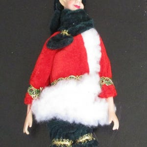 May include: A decorative elf doll wearing a red velvet jacket with white faux fur trim and gold braided accents. The doll has a dark green velvet hat and scarf, dark green pants, and red and white striped leggings with white faux fur cuffs.