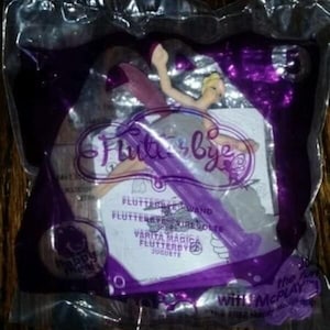 May include: A Flutterbye fairy toy sealed in a clear plastic bag with purple accents. The packaging features the text Flutterbye, Flutterbye Wand, and Happy Meal. The toy is a small, light-colored fairy figure with translucent wings.