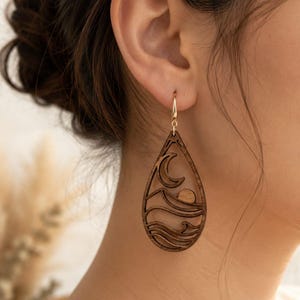May include: A teardrop-shaped wooden earring featuring a laser-cut design of a crescent moon, a sun, and rolling waves. The dark brown wood cutout is suspended from a simple gold-toned metal hook, creating a detailed landscape scene.