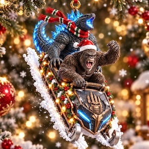 Könnte beinhalten: Ein hängender Weihnachtsbaumschmuck mit Godzilla und King Kong auf einem Schneemobil. Godzilla trägt einen rot-weiß-grün gestreiften Schal, während King Kong eine rote Weihnachtsmannmütze trägt. Das Schneemobil ist mit Stechpalmen und Lichtern verziert.