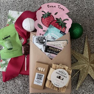 May include: A gift set featuring a brown paper-wrapped package containing a bar of goat milk soap, a small box with a QR code, and various tea packets. Included are a pink strawberry-themed face mask, a green lip mask, and a white bath bomb.