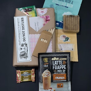 May include: A gift set containing a brown paper-wrapped package with a Do Not Disturb sign, a teal Intensive Neck & Decolletage Mask, a rectangular brown soap bar, a small pouch of bath salts, a Ghirardelli caramel square, and a Barissimo latte mix.