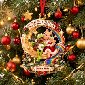 May include: A decorative Christmas ornament featuring Kermit the Frog playing a banjo and Miss Piggy sitting on a golden crescent moon. The text reads WITH YOU, I FOUND MY CHRISTMAS CONNECTION and DAVID EMILY. It includes a rainbow and pond.