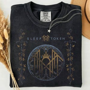 Könnte beinhalten: Ein schwarzes Kurzarm-T-Shirt mit einem Grafikdruck, der den Text Sleep Token, ein zentrales kreisförmiges Emblem mit einem kronenartigen Symbol sowie vertikale, mit Rosen umrankte Schwertmotive auf beiden Seiten zeigt. Eine silberne Sternkette liegt bei.