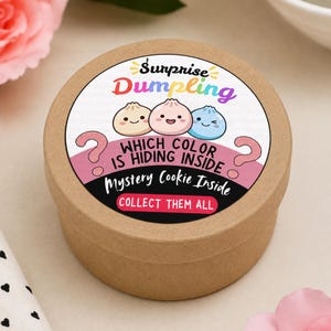 以下が含まれることがあります： 円形の薄茶色の段ボール箱で、上部に丸いラベルが貼られています。ラベルにはベージュ、ピンク、青の3つの餃子のイラストが描かれており、「Surprise Dumpling」、「WHICH COLOR IS HIDING INSIDE」、「Mystery Cookie Inside」、「COLLECT THEM ALL」という文字が記されています。