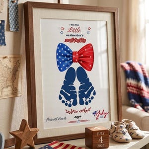 May include: A framed print featuring a red, white, and blue bow tie graphic above two dark blue baby footprints. The text reads I Was This Little on America's 250th Birthday, Made with Love by zaya, and 4th July 2026. The frame is dark wood.
