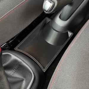 May include: A black plastic car centre console organiser tray with a rectangular storage compartment and a curved cutout section designed to fit securely around the handbrake lever base, providing additional storage space between the vehicle seats.