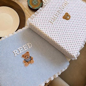 May include: Two square fabric covers with white lace trim. One cover is light blue with white vertical stripes, featuring the embroidered name REED and a brown teddy bear patch. The other is white with a small floral print, the name EMERSON, and a bear face.