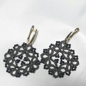 Puede incluir: Un par de pendientes colgantes con motivos circulares negros similares al encaje, adornados con pequeñas cuentas oscuras. Los pendientes están sujetos a cierres de palanca de metal dorado pulido, creando un contraste entre los intrincados patrones oscuros y el metal.