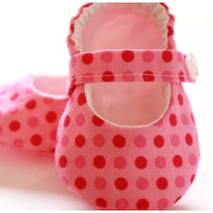 May include: A pair of baby shoes in shades of pink with a polka dot pattern. The shoes have a Mary Jane style with a strap and button closure. The interior lining is white.