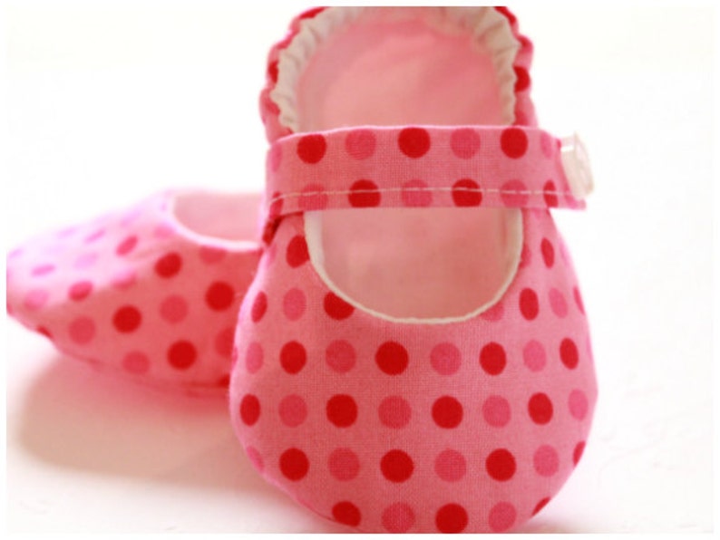 May include: A pair of baby shoes in shades of pink with a polka dot pattern. The shoes have a Mary Jane style with a strap and button closure. The interior lining is white.