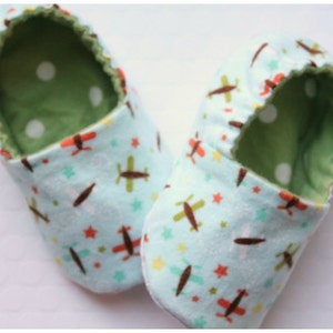 May include: A pair of baby booties with a light blue airplane pattern. The booties have a green interior with white polka dots. The exterior features brown, red, and green airplanes and stars.