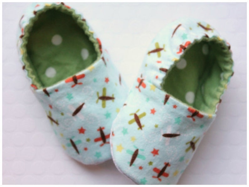 May include: A pair of baby booties with a light blue airplane pattern. The booties have a green interior with white polka dots. The exterior features brown, red, and green airplanes and stars.