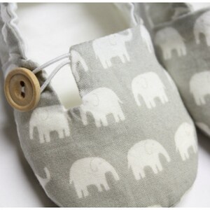 May include: A pair of grey baby shoes featuring a white elephant pattern. The shoes have a wooden button fastening and elastic cord. The interior is white.