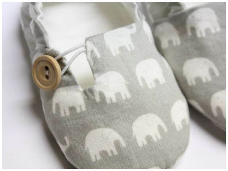 May include: A pair of grey baby shoes featuring a white elephant pattern. The shoes have a wooden button fastening and elastic cord. The interior is white.