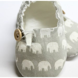 May include: Baby shoes featuring a grey elephant pattern. The shoes have a light grey interior and a wooden button fastening. The fabric is soft, and the shoes appear to be handmade.