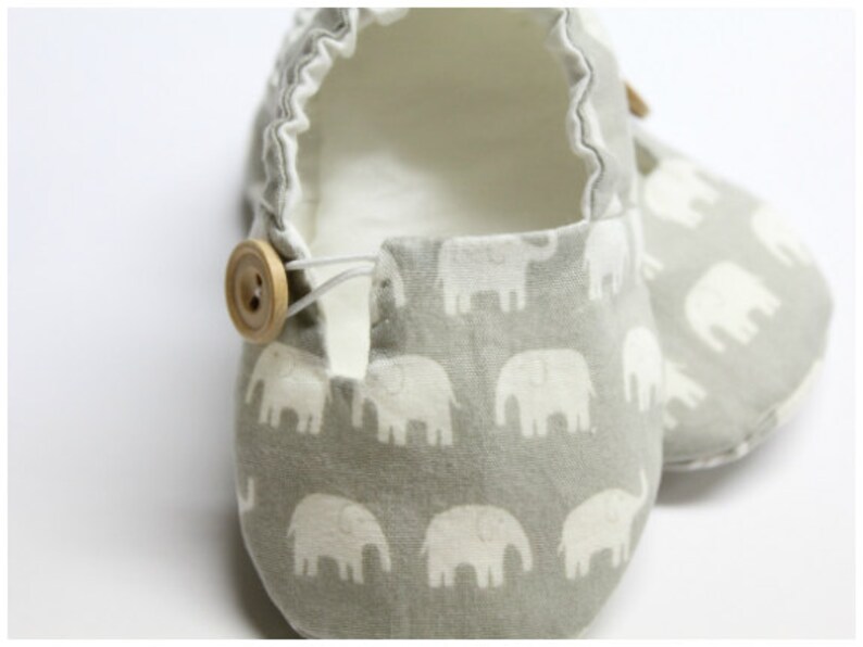 May include: Baby shoes featuring a grey elephant pattern. The shoes have a light grey interior and a wooden button fastening. The fabric is soft, and the shoes appear to be handmade.
