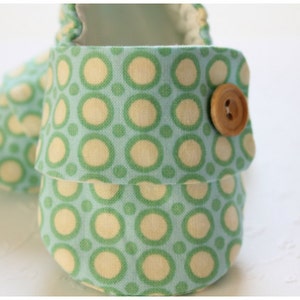 May include: Close-up of a pair of baby shoes. The shoes are made of a light blue fabric with a repeating pattern of green and cream-colored circles. A wooden button secures the flap. The shoes are designed for infants.