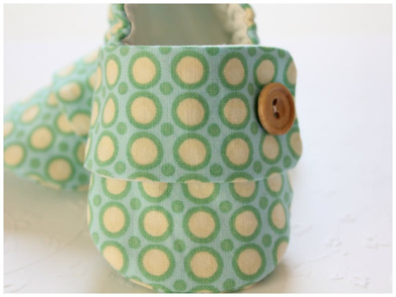 May include: Close-up of a pair of baby shoes. The shoes are made of a light blue fabric with a repeating pattern of green and cream-colored circles. A wooden button secures the flap. The shoes are designed for infants.