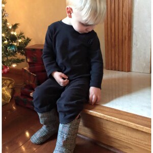 May include: A young child wearing a black long-sleeved shirt and black pants, seated on a wooden ledge. The child is wearing plaid boots with red and black accents. A Christmas tree is visible in the background.