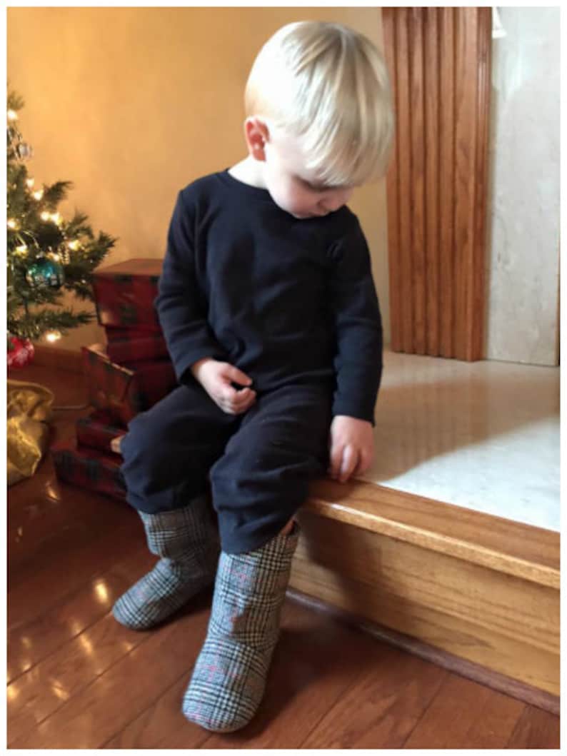 May include: A young child wearing a black long-sleeved shirt and black pants, seated on a wooden ledge. The child is wearing plaid boots with red and black accents. A Christmas tree is visible in the background.