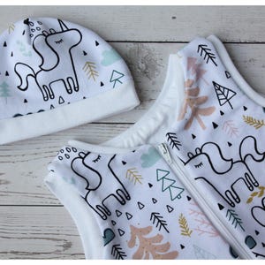 May include: A white baby sleep sack and matching hat set. The set features a whimsical unicorn design with black outlines, along with trees and geometric shapes in pastel colors. The sleep sack has a zipper closure.