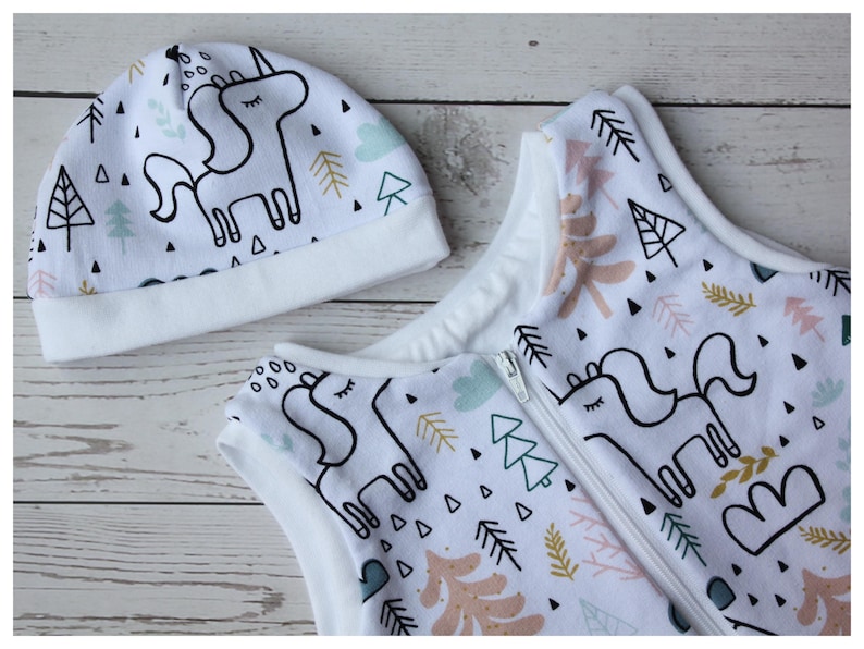 May include: A white baby sleep sack and matching hat set. The set features a whimsical unicorn design with black outlines, along with trees and geometric shapes in pastel colors. The sleep sack has a zipper closure.